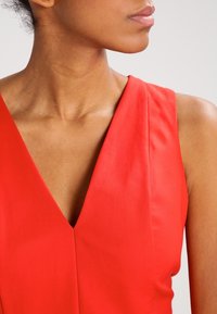Cuello y hombro de mujer en una blusa roja sin mangas con escote en V, contra un fondo gris claro liso.