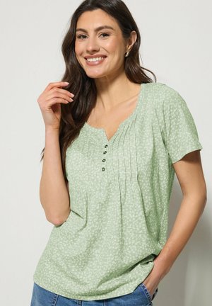 Vrouw die glimlacht, gekleed in een lichtgroene, patroonrijke top met korte mouwen, knopen en plooien, poserend met één hand bij haar gezicht en de andere in haar zak.