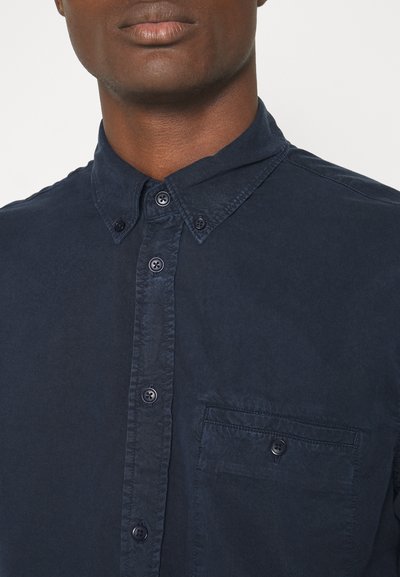 Camicetta blu navy con colletto a punta, cuciture visibili e taschino sul petto. Realizzata in tessuto liscio con due bottoni al colletto.