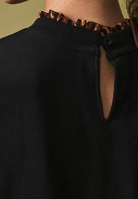 Vêtement noir avec un col brun texturé et volanté. Dispose d'une fermeture à boutons et d'une ouverture en forme de goutte d'eau au dos. Texture du tissu douce.