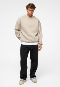 Beige oversized sweatshirt met ronde hals, gelaagd over een wit shirt, gecombineerd met losse zwarte jeans en grijze sneakers, met zilveren kettingen.