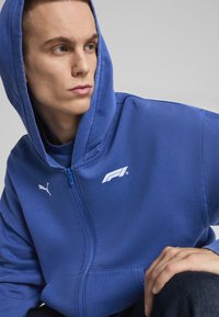 Plava dukserica na zip s mekanom tkaninom, koja ima kengurucu džep, kapuljaču i bijele logotipe: Puma emblem i F1 grafiku na prsima.