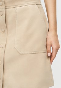Μπεζ φούστα με κουμπιά στο μπροστινό μέρος, μεγάλο λοξό μπροστινό τσέπη, ορατές ραφές και ένα χέρι που ακουμπά δίπλα στην τσέπη.