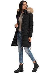 Zwarte pufferjas met een capuchon van nep-bont, ritssluiting en drukknoopsluiting, schuine zakken en bruine leren accenten; gedragen met blauwe jeans en zwarte laarzen.