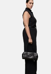 Bolso de mano de cuero negro con acabado texturizado, asas dobles y cierre de broche. El modelo lleva una blusa sin mangas negra y pantalones de sastre.