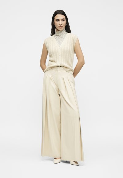 Γυναίκα με μπεζ αμάνικο γιλέκο με κουμπιά πάνω από ριγέ ζιβάγκο, φαρδιά πλισέ κρεμ παντελόνια και μυτερά τακούνια, στέκεται με τα χέρια πίσω.