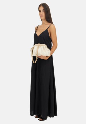 Femme portant une longue robe noire sans manches tenant une pochette beige avec une bandoulière, debout sur un fond clair uni.