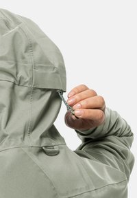 Giacca a vento verde chiaro con tessuto texturizzato. Una mano regola il cordino al collo. Le caratteristiche includono una zip e linguette laterali.