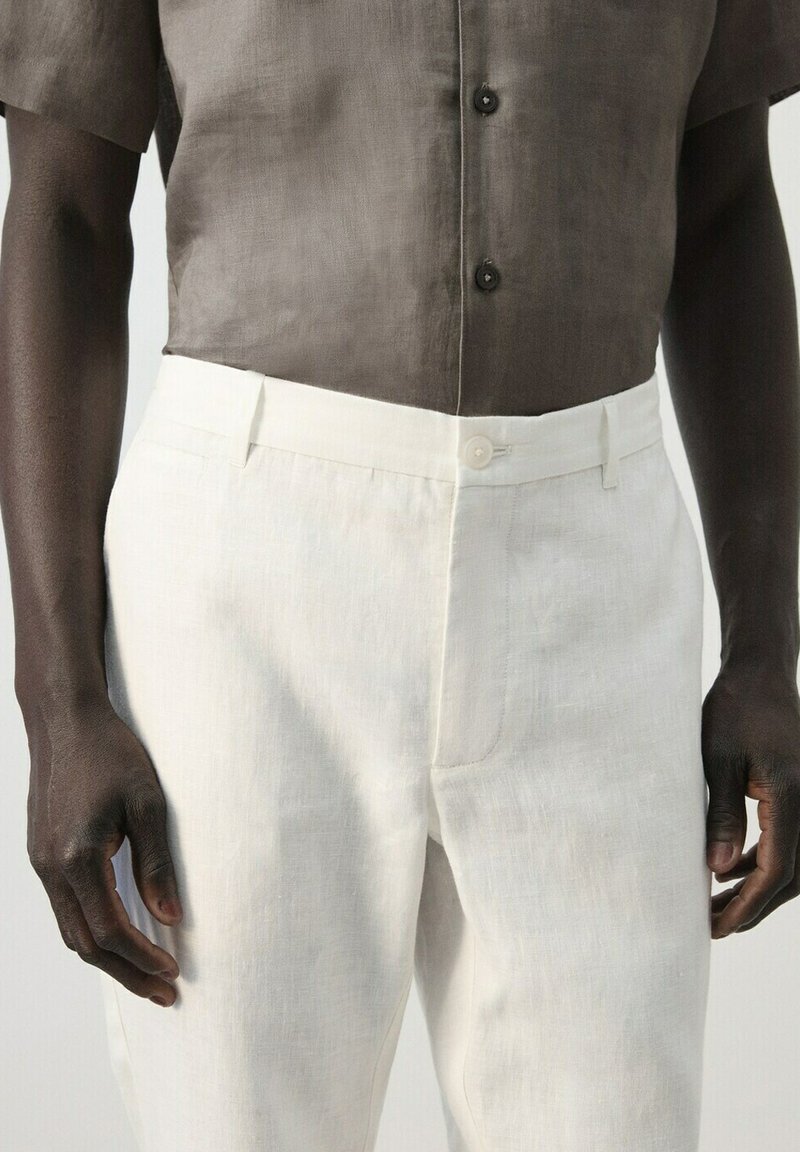 Homme portant une chemise à manches courtes marron clair rentrée dans un pantalon en lin blanc taille haute avec fermeture à boutons.