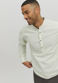 Camisa de manga larga verde claro con cuello de botón, que presenta botones de madera y un dobladillo ligeramente curvado. Tela sencilla y texturizada.