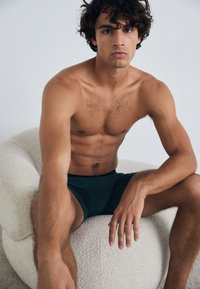 Zöld boxer alsónadrág egy férfi modellen, aki egy textúrázott krémszínű széken ül, izmos felépítéssel és göndör hajjal, egy egyszerű háttér előtt.