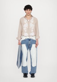Camicia beige con bottoni e ricami floreali bianchi, abbinata a jeans oversize con toppe blu e bianche. Scarpe nere completano il look.