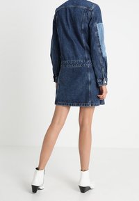 Denim dress with contrast denim panel, tailored fit, and cinched waist. Features long sleeves and a straight hem, paired with white ankle boots.