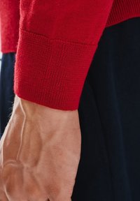 Czerwony sweter dziany z prążkowanym mankietem, prezentujący gładkie i miękkie wykończenie. Skóra częściowo widoczna pod mankietem.