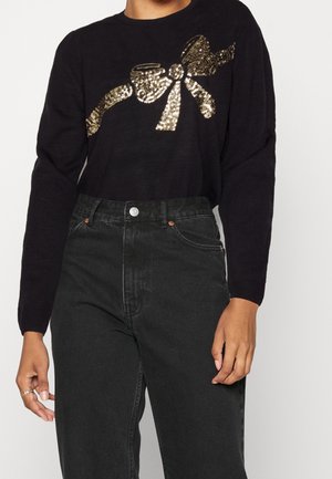Schwarzer Pullover mit langen Ärmeln und einem großen goldenen Pailletten-Schleifenmotiv über der Brust, kombiniert mit hoch taillierten schwarzen Jeans.