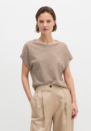 Kobieta o krótkich brązowych włosach, ubrana w beżową i białą pasiastą bluzkę z krótkim rękawem oraz beżowe spodnie z wysokim stanem, z rękami w kieszeniach.