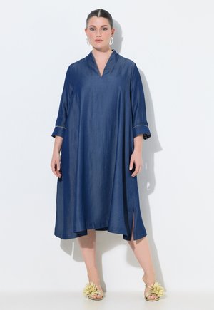 Kobieta ubrana w luźną, granatową sukienkę do kolan z rękawami do połowy przedramienia i rozcięciem, w połączeniu z sandałami ozdobionymi dużymi kwiatowymi dekoracjami.