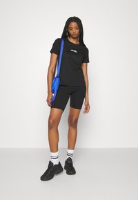 Fekete, rövid ujjú póló fehér logóval, fekete kerékpáros sorttal párosítva. Kiegészítők közé tartozik egy kék crossbody táska és fekete sportcipő.