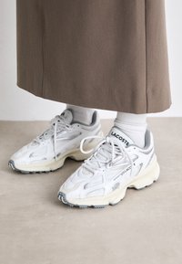 Hvite joggesko med teksturert mesh-overdel, grå accenter, og en klumpete såle. Snøringer og merke er synlige på tungen. Brukt med hvite sokker.