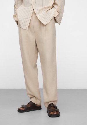 Pantaloni beige a taglio largo abbinati a sandali scuri marrone con punta aperta e camicia a righe chiara con bottoni.