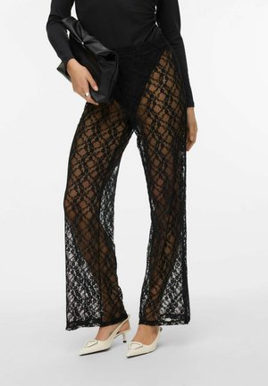 Pantalons en dentelle noire à motif floral, en tissu transparent, avec des jambes larges. Associés à un sac noir et des chaussures à talons kitten blanches.