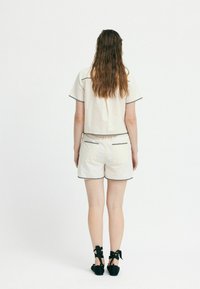 Completo crema con top e pantaloni corti, arricchito da dettagli di cucitura nera. Il top ha maniche corte e i pantaloni corti presentano tasche posteriori. Cinturini neri alla caviglia.