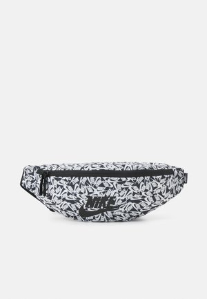 Riñonera negra y blanca de Nike con patrón repetido de logotipo y un bolsillo frontal con cremallera sobre un fondo liso.