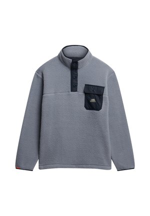 Sudadera de felpa gris con media cremallera, cuello alto, bolsillo en el pecho en color azul marino y parche de logo de montaña. Textura suave y ajuste relajado.