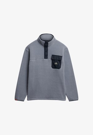 Szary polarowy narzut z pół zamkiem, wysokim kołnierzem, kieszenią na piersi w kolorze granatowym oraz naszywką z logo gór. Miękka struktura i luźny krój.