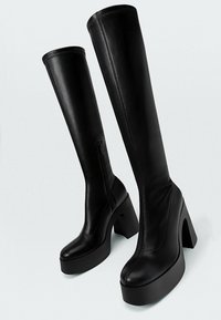 Bottes en cuir noir jusqu'au genou avec une plateforme épaisse et un talon bloc incurvé. Dotées d'une fermeture éclair sur le côté et d'une texture lisse.