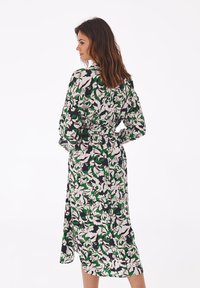 Langärmeliges Blumenkleid mit schwarzem Hintergrund, das grüne und weiße Blattmuster zeigt, eine gebundene Taille hat und lange Ärmel besitzt.