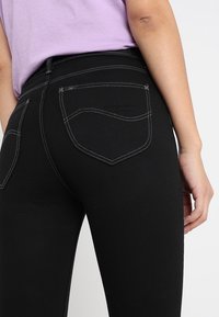 Persona vestida con pantalones negros ajustados con costuras blancas y una camisa lavanda metida por dentro, de pie contra un fondo claro y liso.
