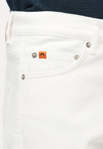 Τσέπη από λευκό τζιν με μεταλλικά καρφιά και μικρή πορτοκαλί ετικέτα λογότυπου από ύφασμα, φορεμένη με σκούρο τοπ.