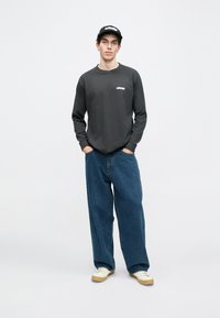 Camisa cinza de mangas longas com um pequeno logótipo, combinada com jeans azul de corte solto e ténis brancos com solas bege. Boné preto usado.