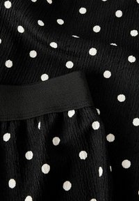 Crni materijal s bijelim točkicama, s teksturiranom površinom. Na vrhu je vidljiv elastični pojas.