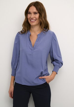 Lichtblauwe blouse met een V-hals, driekwart pofmouwen en gestructureerde strepen. Gecombineerd met donkere broek en een zakdetail.