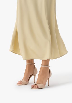 Beżowe sandały na wysokim obcasie z odkrytymi palcami i paskami wokół kostki, noszone z lekką, kremową, lejącą się spódnicą na białym tle.