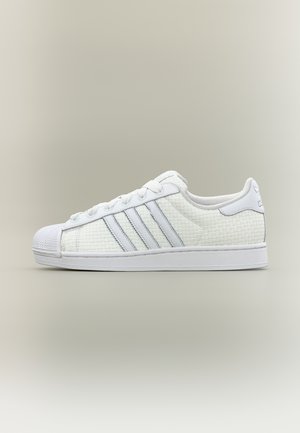 White low-top sneaker with textured woven upper, rubber shell toe, lace-up closure, and three stripes on the side.