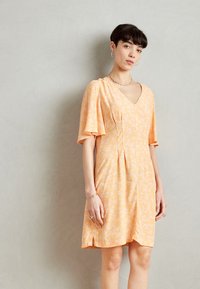 Robe à manches courtes en tissu orange doux avec des motifs floraux blancs. Design fluide avec des plis subtils et des fentes sur les côtés pour le mouvement.