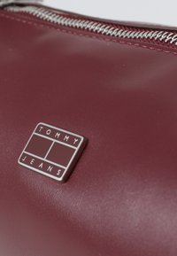 Punainen synteettinen pussi, jossa on hopeaketju, ja jossa on suorakulmainen metallinen logolevy, jossa on kohokuvioituna "TOMMY JEANS". Suora pinta.