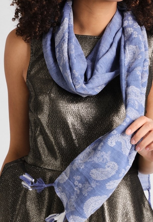 Femme portant une robe métallisée sans manches, tenant un foulard bleu à motif cachemire avec des glands autour du cou.