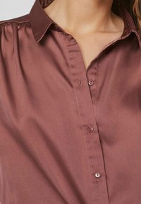 Kastanjefärgad satinblus med krage, som har en knappslå och subtil plissering. Slät textur med en mjuk lyster.