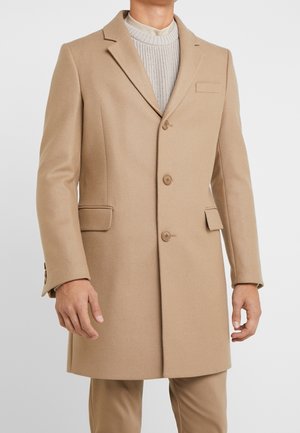 Mężczyzna w beżowym, jednorzędowym płaszczu, na jasnym dzianinowym swetrze, stoi z ramionami swobodnie opuszczonymi wzdłuż ciała.