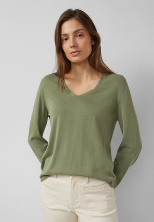 Donna con capelli castano chiaro che indossa un maglione a V verde oliva e pantaloni beige chiaro davanti a uno sfondo grigio uniforme.