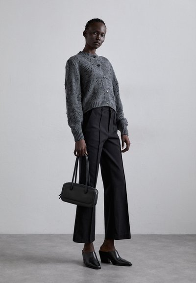 Grau gestrickte Strickjacke mit Knöpfen, weite schwarze Hose und schwarze hohe Mules. Trägt eine kleine rechteckige schwarze Handtasche.