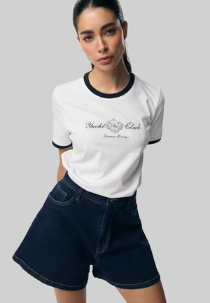 Kobieta z ciemnymi, spiętymi włosami, ubrana w białą koszulkę Yacht Club z czarną lamówką oraz ciemnoniebieskie szerokie szorty dżinsowe, stojąca na szarym tle.