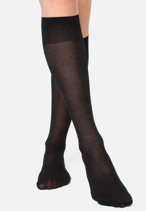 Legs wearing sheer black knee-high stockings with a solid band at the top, standing on a light gray background.