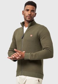 Chaqueta de cremallera verde oliva con tejido texturizado acolchado, puños y dobladillo acanalados, que presenta un pequeño logo circular en el pecho.