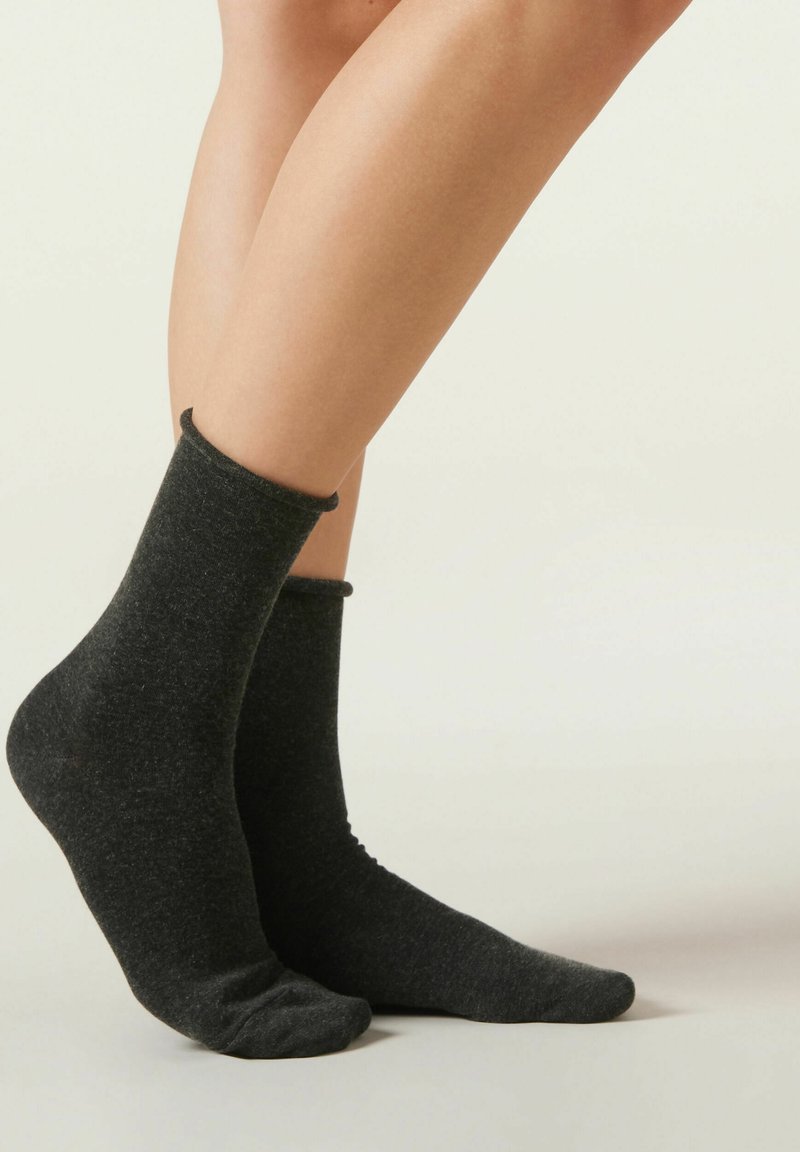 Piernas con calcetines gris oscuro hasta la mitad de la pantorrilla, de pie sobre una superficie lisa y clara con un fondo claro y uniforme.