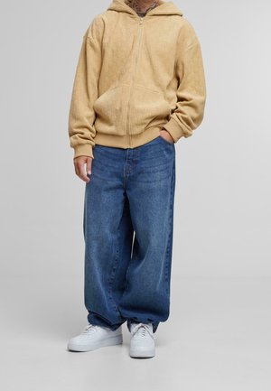 Beige Kapuzenpullover mit Reißverschluss und strukturiertem Muster, getragen zu lockeren blauen Jeans und weißen Turnschuhen. Der Hoodie hat eine Kapuze mit Kordelzug.